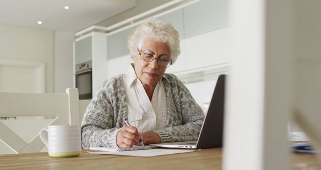 Elderly woman working with a laptop and writing notes at a table in a modern home environment. Ideal for concepts related to seniors working from home, remote work, aging and technology, productivity among the elderly, and active senior lifestyle.