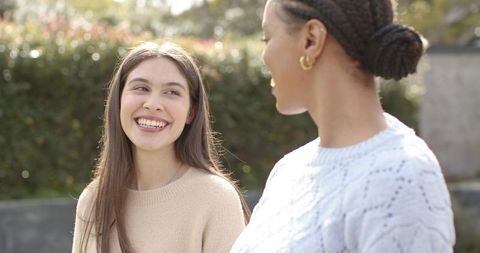 Smiling friends chatting outdoors wearing knit sweaters and gold hoop earrings in sunlight
