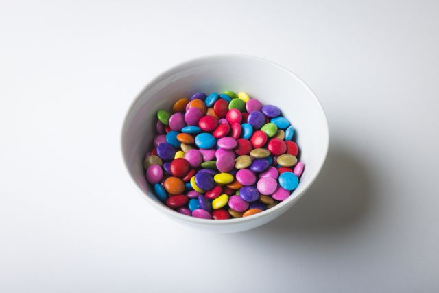 High angle view of a bowl filled with vibrant, multi-colored chocolate candies against a plain white background. Ideal for use in advertisements, blogs, or articles related to sweets, desserts, and snacks. Perfect for illustrating concepts of indulgence, treats, and unhealthy eating habits.