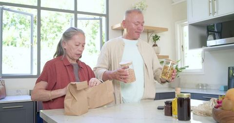 Senior couple unpacking groceries and organizing pantry jars on bright marble kitchen island