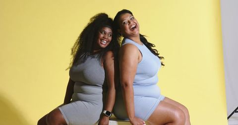 Plus-size diverse women laughing back-to-back wearing sporty dresses on yellow backdrop