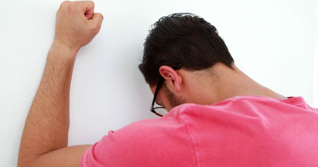 A young Caucasian man appears frustrated or upset, leaning his forehead against a wall with his fist raised. His body language suggests stress, disappointment, or exhaustion, emphasizing the emotional weight he may be experiencing.
