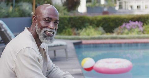 African american senior man sitting by pool smiling, relaxing with float and beach ball