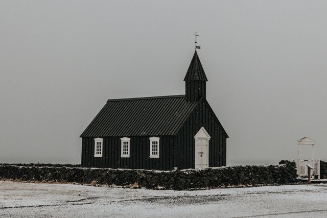 Traditional black church stands alone against a snowy, overcast backdrop. The rustic structure, surrounded by a low stone wall, evokes a sense of solitude and peace. Perfect for use in travel blogs about Iceland, religious settings, winter-themed projects, and showcasing historic architecture.