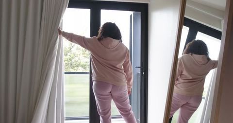 Curly-haired woman standing at patio door pulling curtain wearing pink hoodie and sweatpants