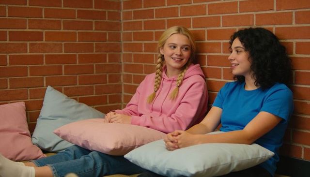 Two teens chatting on cozy lounge cushions with pastel pillows and brick wall backdrop