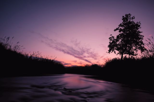 This tranquil scene depicts a river flowing under a dramatic twilight sky. The silhouette of a lone tree stands against the backdrop of a purple and orange sky with streaky clouds. Great for use in relaxation or calming products, meditation visuals, nature articles, and motivational or inspirational social media posts.