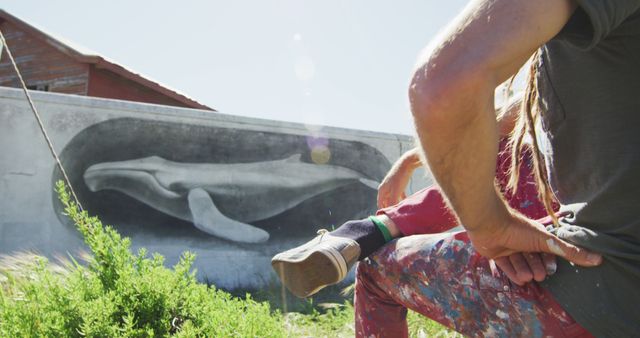 Street artist taking a break in front of a large whale mural. Ideal for illustrating concepts of urban art, creativity, and the artistic process. Useful for blogs, articles, and promotional materials related to street art and muralists.