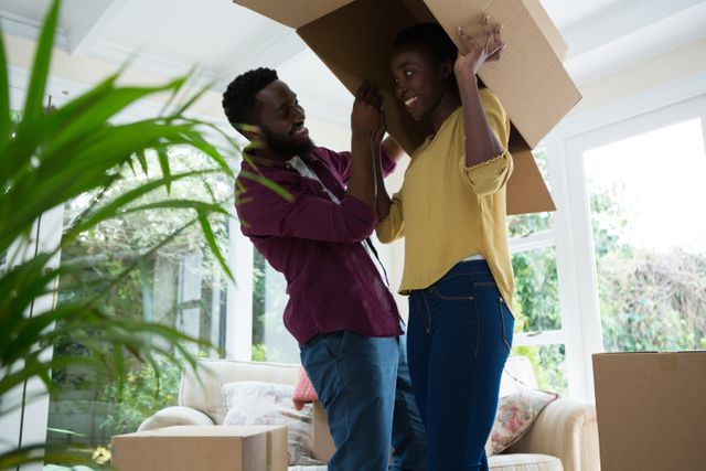 This image shows a joyful couple playing with a cardboard box while moving into their new house. They are smiling and enjoying the moment, surrounded by unpacked boxes in a bright living room. This image can be used for themes related to moving, new beginnings, family bonding, and home life.