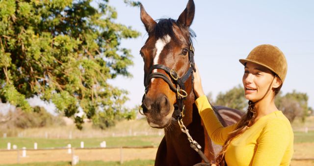 A woman in a yellow shirt and riding helmet enjoys time with a horse outdoors on a sunny day. Suitable for concepts related to leisure, hobbies, nature, and equestrian sports. Ideal for promotional materials for horse riding schools, animal bonding experiences, and outdoor recreational activities.