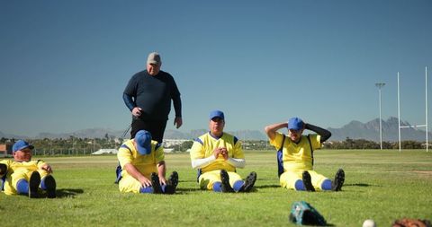 Community baseball team sitting during pregame warmup while coach observing field drill