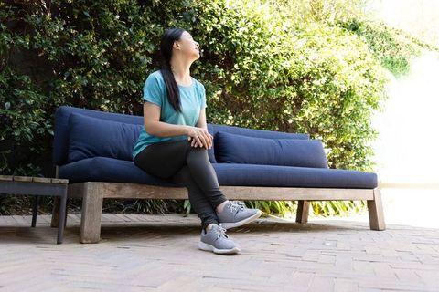 Asian woman sitting on outdoor wooden bench with blue cushions, looking up, relaxing