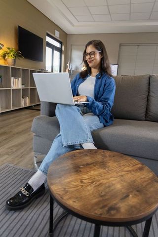 Young woman working on laptop on sofa in modern living room, casual remote work lifestyle