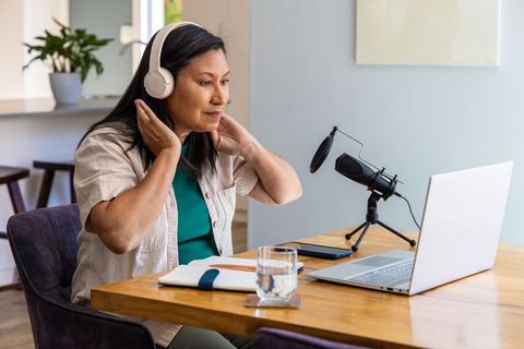 Mature asian woman adjusting headphones while recording podcast at home studio desk