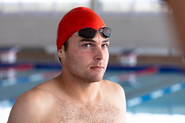 Portrait of Caucasian male swimmer wearing swimming goggles and red swimming cap looking at camera in a swimming pool before competing. Sports athletic competition.