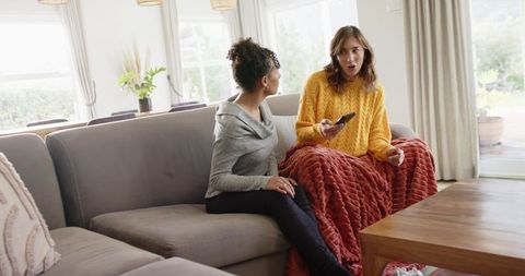 Two friends chatting on sofa wrapped in knit blanket holding remote in bright cozy living room