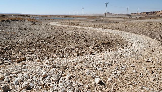 Snaking pale gravel ribbon crossing arid plains with power lines, pond and rocky scrub