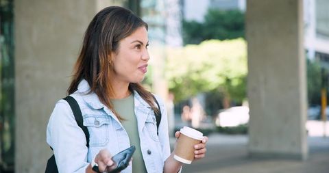 Young woman holding coffee and smartphone smiling on urban plaza wearing denim jacket