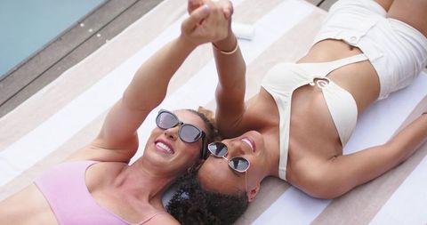 Smiling diverse women lying on striped towel at poolside wearing sunglasses and swimwear