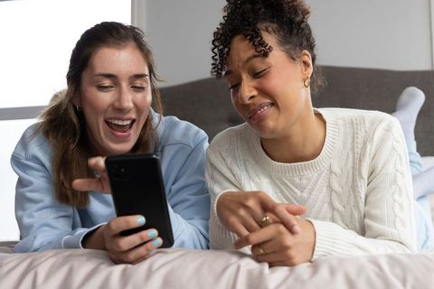 Young women laughing, sharing smartphone on bed cozy casual lifestyle moment