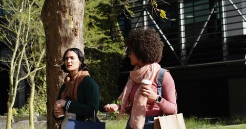 Young women walking and talking in sunlit campus courtyard holding coffee cups wearing scarves