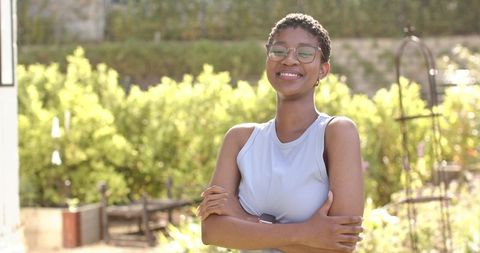 Smiling african woman in activewear with glasses and smartwatch, arms crossed in sunny backyard