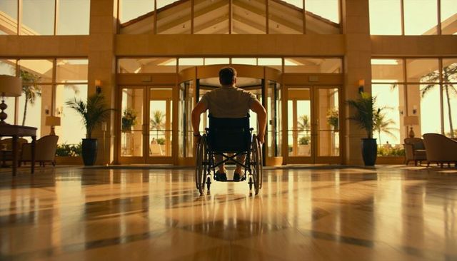 Man in wheelchair rolling toward revolving door in sunlit resort lobby atrium