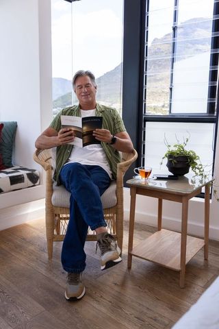 Mature man relaxing with book in sunlit rattan chair by mountain view window