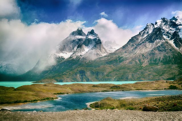 This stock photo showcases the breathtaking Andes Mountains with snow-capped peaks surrounded by turquoise lakes and clouds drifting above. It is perfect for use in travel brochures, nature documentaries, environmental websites, or inspirational posters emphasizing the beauty of natural landscapes.