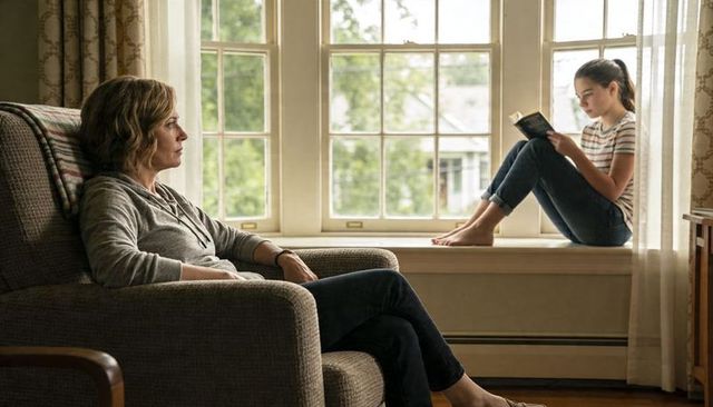 Mother and teenage daughter sharing quiet moment, daughter reading on windowsill in sunlit living ro