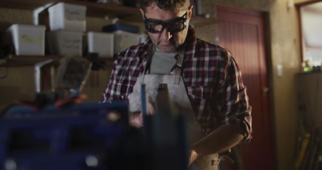 Middle-aged man in a workshop environment, concentrating on his task. He wears safety glasses and a plaid shirt, ensuring protection while working with tools and machinery. Ideal for depicting themes related to craftsmanship, manual labor, and safety in industrial settings. Useful for articles on DIY projects, industrial safety, or craftsmanship expertise.