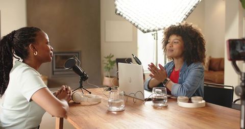 Diverse women recording conversational podcast in cozy home studio with softbox lighting