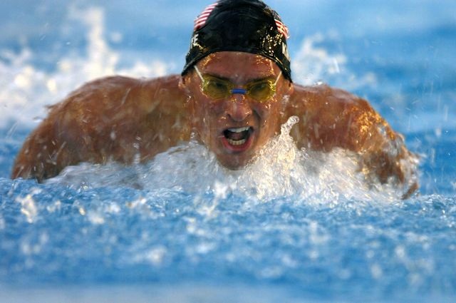 Competitive swimmer wearing goggles and swim cap is performing butterfly stroke in a swimming pool. The image captures the intensity and focus required in competitive swimming. Ideal for articles or advertisements related to swimming, athletics, sports training, swim gear promotions, and motivational content about dedication and determination.