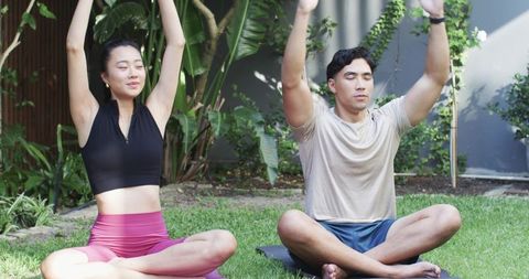 Couple practicing yoga in backyard garden, cross-legged meditation on mats, tropical foliage