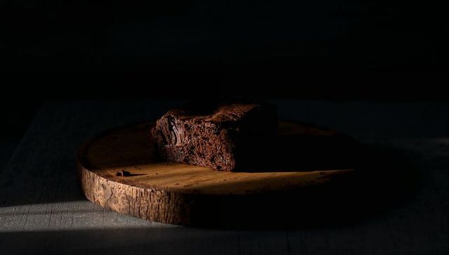 Dense Dark Chocolate Brownie Resting on Round Wooden Board Casting Dramatic Shadow