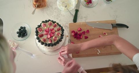 Friends decorating frosted layer cake on countertop with figs, strawberries and blueberries