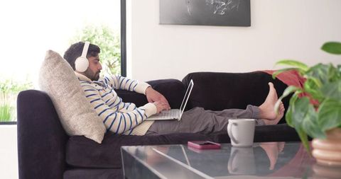 Indian man in striped sweater reclining on sofa working on laptop with headphones barefoot