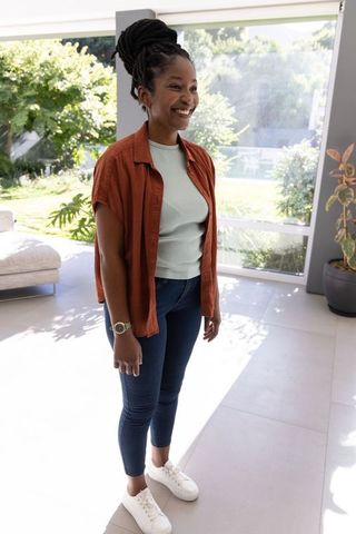 Smiling young black woman standing by floor-to-ceiling window in sunlit modern living room