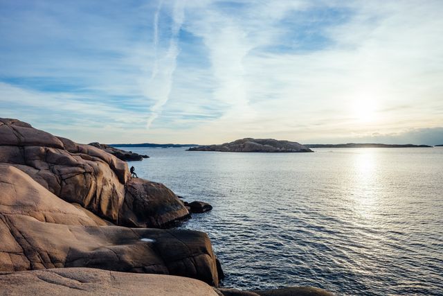 This photo features a person standing on a rocky shore, looking out over the ocean at sunset. The sky is partly cloudy with the sun casting a warm glow on the water and rocks. Ideal for websites or brochures promoting travel, adventure, and nature activities.