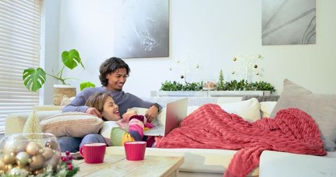 Asian couple relaxing on sectional couch with laptop, festive holiday decor, cozy throw