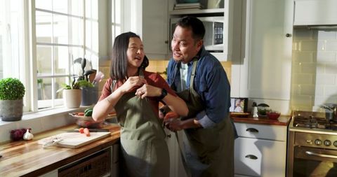 Asian Father and Teen Daughter Cooking Together in Sunlit Kitchen Wearing Green Aprons