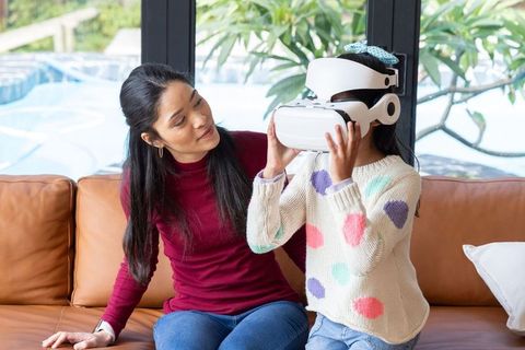 Asian mother watching daughter adjusting white vr headset on couch, family technology bonding