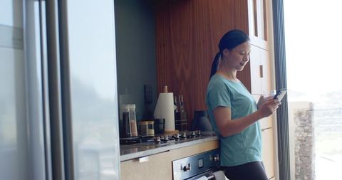 Asian woman leaning on modern kitchen counter using smartphone with sunlit terrace view