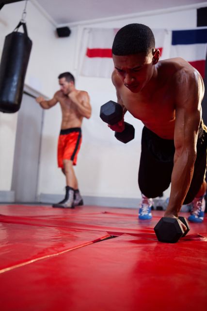 Two men are engaged in an intense workout session in a fitness studio. One man is performing a dumbbell exercise on a red mat, showcasing his strength and determination. The other man is seen in the background, also focused on his fitness routine. This image is ideal for use in fitness-related content, gym promotions, health and wellness articles, and sports training materials.