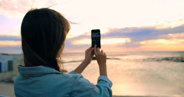 Woman holding smartphone, capturing beautiful sunset over ocean. Ideal for travel blogs, vacation ads, outdoor activities, tech usage in everyday life themes, nature appreciation.