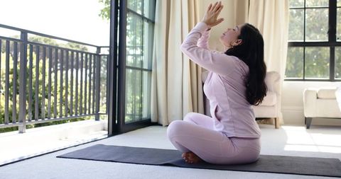 Woman practicing seated meditation in pastel pink loungewear on yoga mat by sunlit balcony