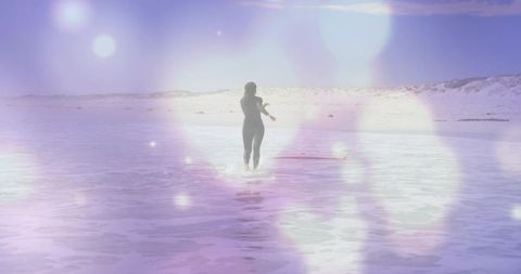 Woman walking and wading across shallow sea toward dunes with surfboard, dreamy lens flare