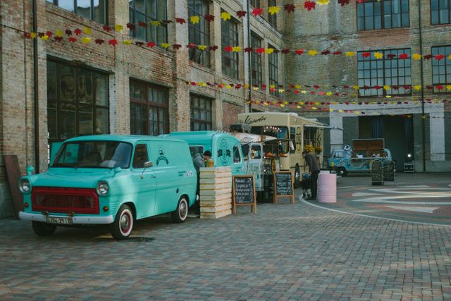 Vintage food trucks are parked in an open, urban industrial area with old brick buildings and colorful decorations overhead. Ideal for articles or advertisements about street food festivals, urban markets, or retro-themed events.