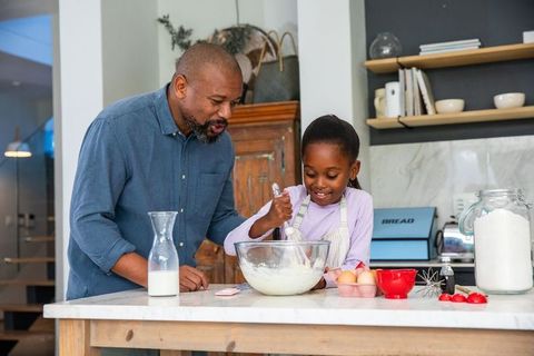 African american father guiding daughter mixing batter at kitchen island, smiling while baking toget