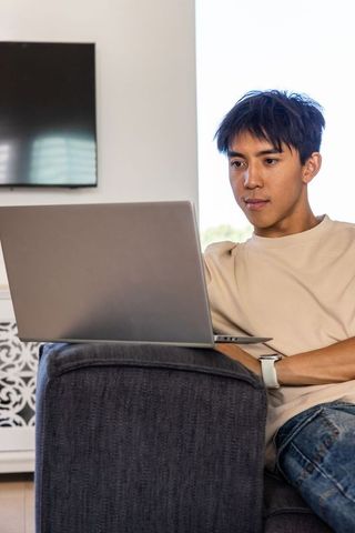 Asian man working on laptop on gray couch in modern living room, remote work focus